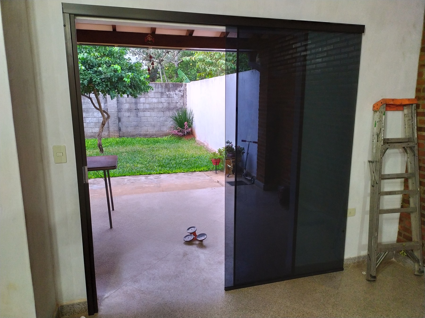 Puerta corrediza de vidrio gris sin marco abierta desde el interior hacia el patio · MÁS VIDRIOS