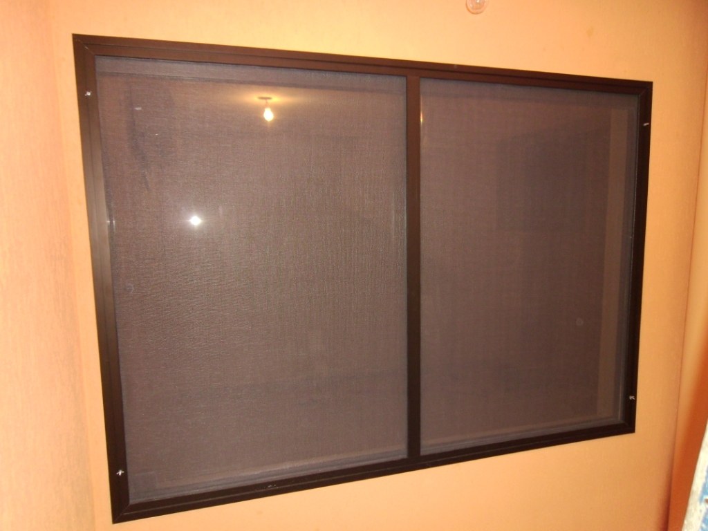 Mosquitero de dos paños con marco de aluminio marrón sobre una ventana, en pared naranja · MÁS VIDRIOS