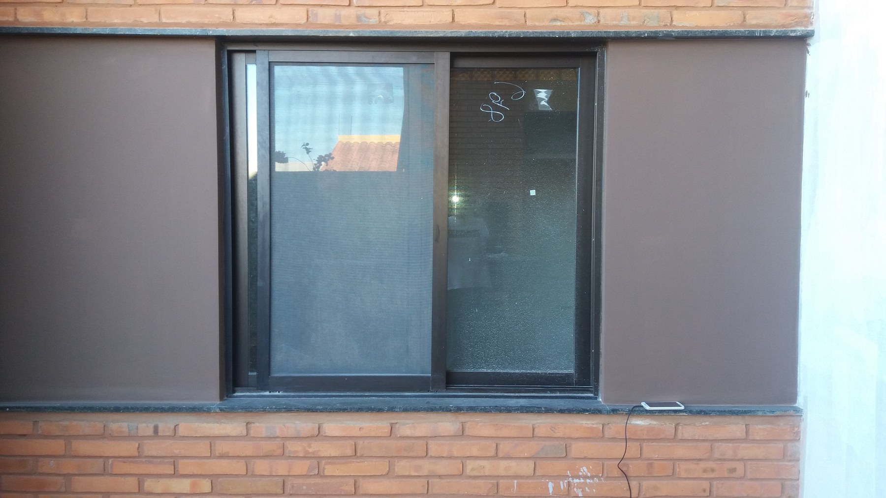 Ventana corrediza de aluminio oscuro con mosquitero en pared revocada con zócalo de ladrillo · MÁS VIDRIOS